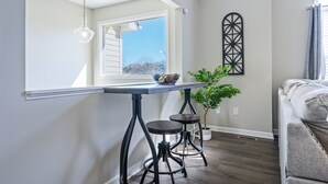 A modern kitchen with a long counter, two stools, a hanging light, a window with blinds, and a decorative wall piece.