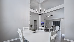Un vaste espace dédié à la salle à manger, avec une table et des chaises blanches, un lustre et un ventilateur de plafond.