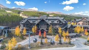 Un chalet à plusieurs étages, avec un télésiège devant, entouré d’arbres aux couleurs automnales et avec des montagnes en arrière-plan.