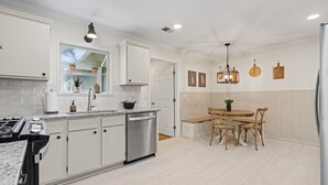 A modern kitchen with white cabinetry, a stainless steel dishwasher, and a granite countertop. There is a dining area with a wooden table and chairs.