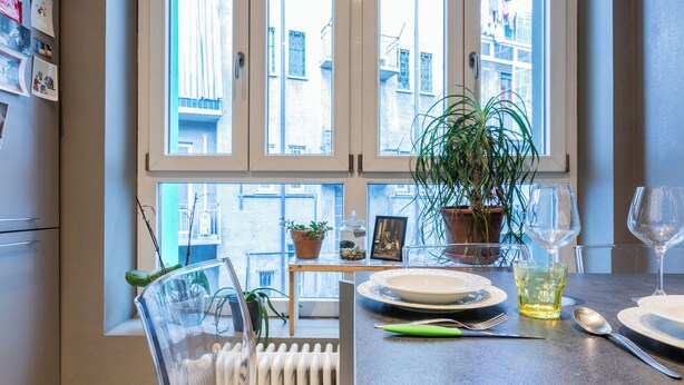 Ett kök med fönsterutsikt, ett matbord dukat med tallrikar, glas och bestick, och en krukväxt på fönsterbrädan.