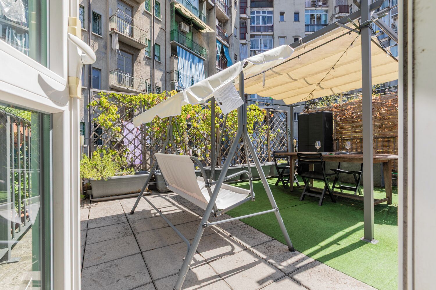 An outdoor patio with a swing set, a table with chairs, and a green artificial turf area.