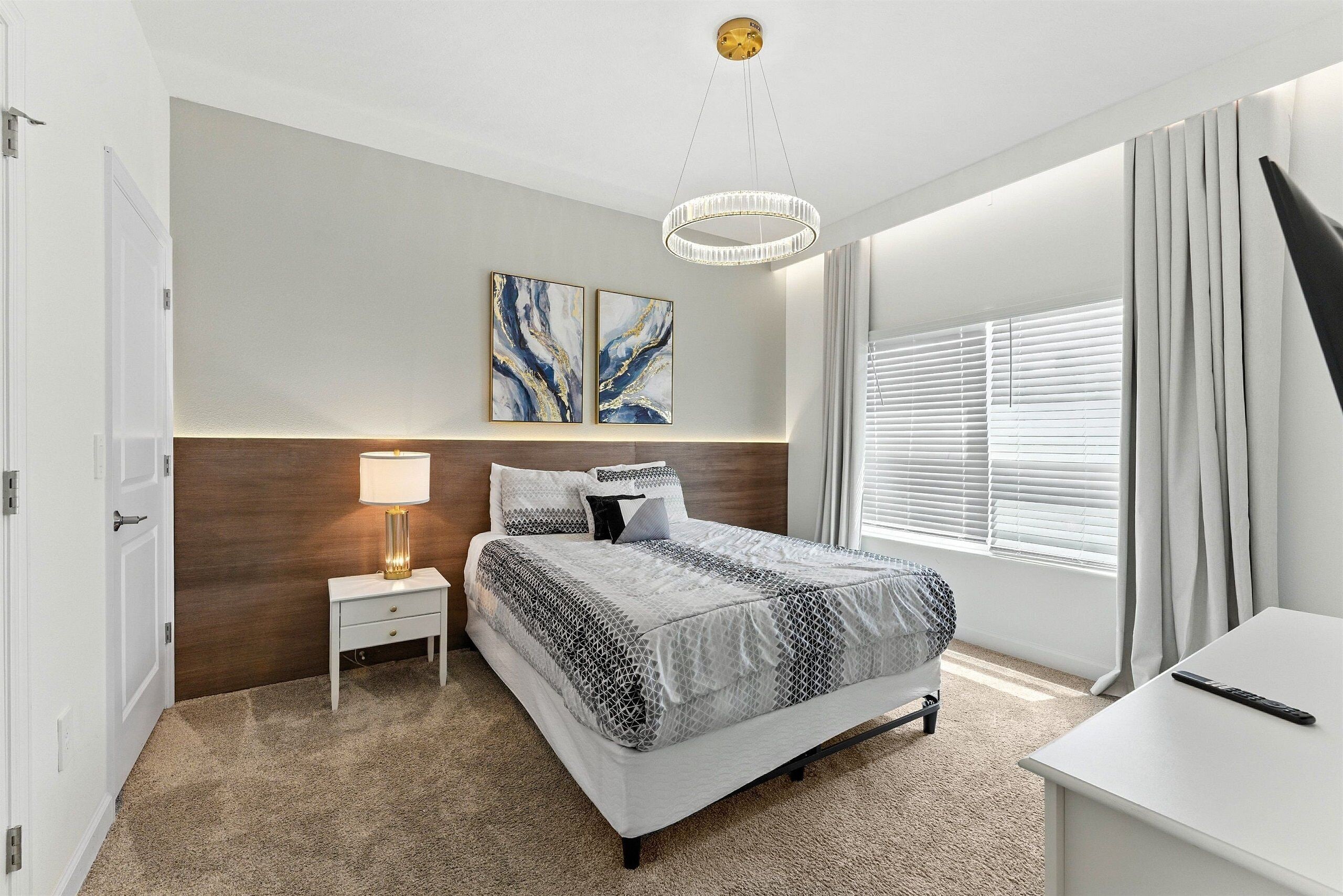 Een slaapkamer met een bed, nachtkastje, lamp, kunstwerk en een raam met gordijnen.