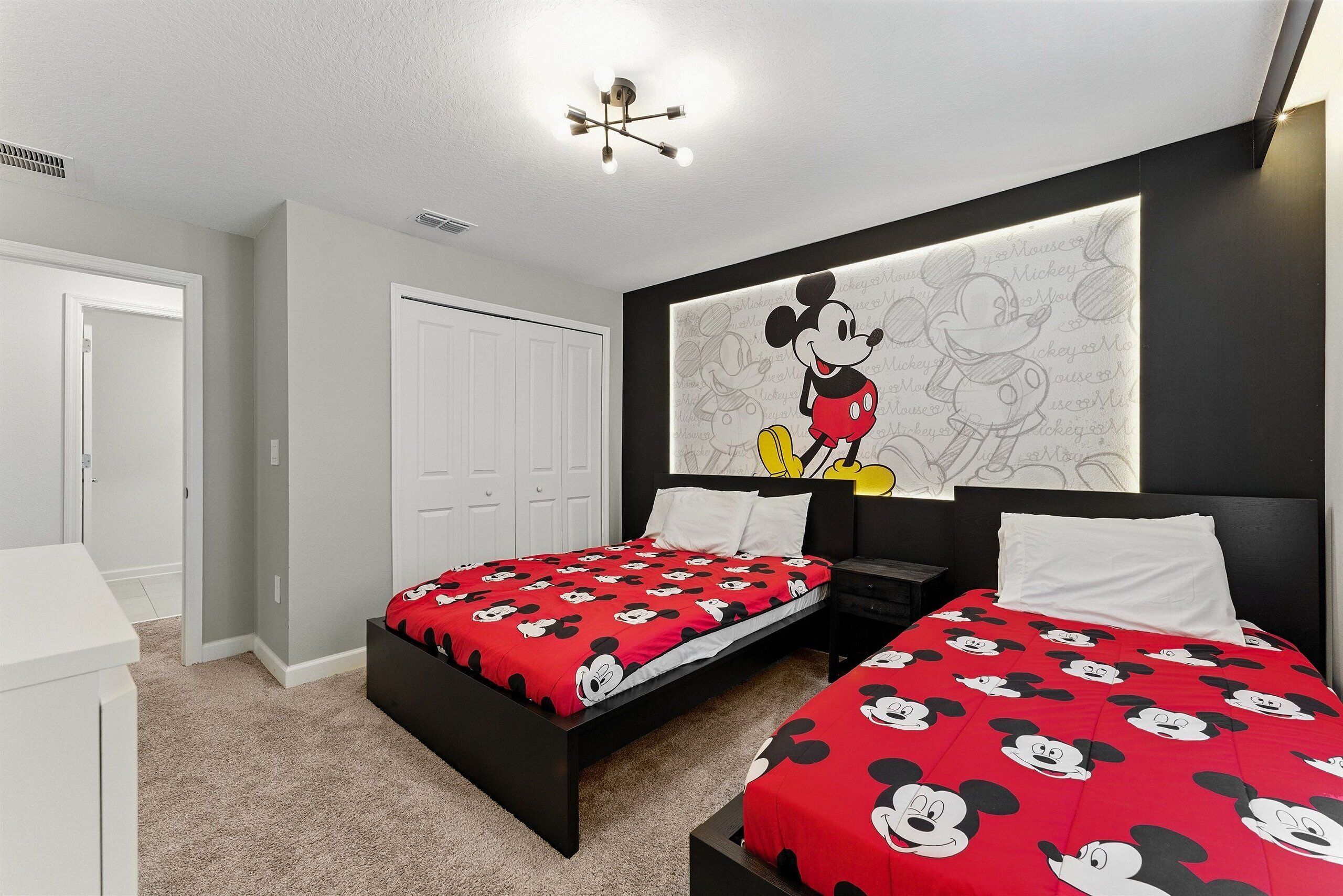 A bedroom with two beds, a large Mickey Mouse mural on the wall, and a ceiling light fixture.