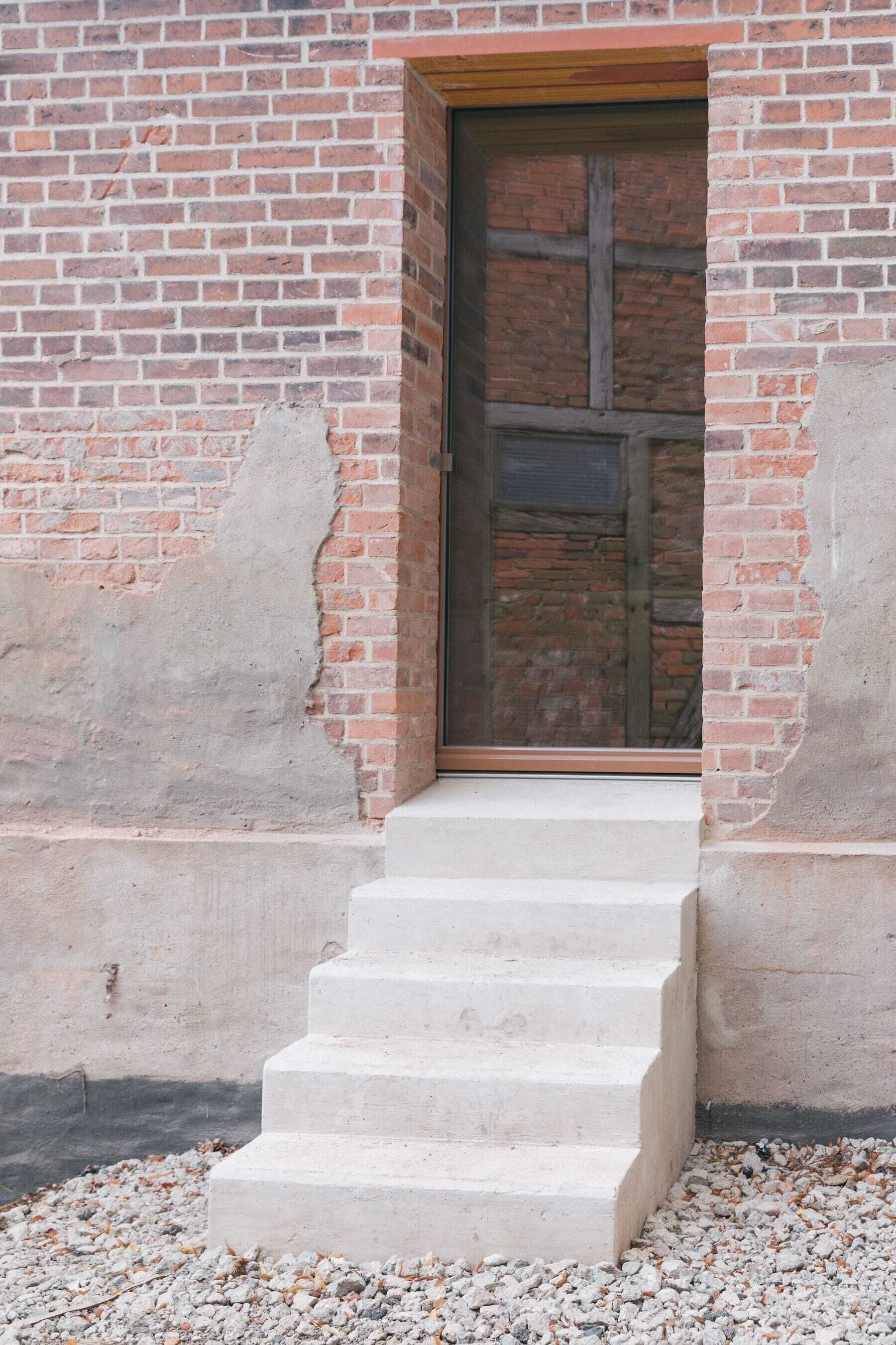 Una escala de formigó que porta a una porta de fusta, amb un mur de maó.