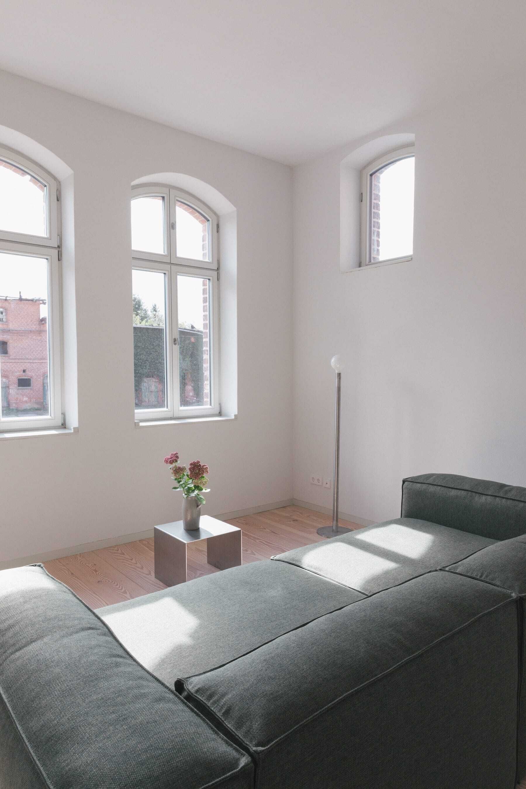 Un salon moderne avec un canapé gris, une petite table sur laquelle est posé un vase de fleurs et trois fenêtres qui laissent entrer la lumière naturelle.