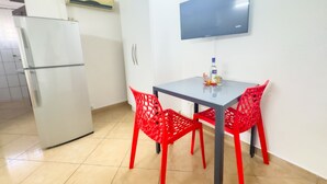 A small kitchen with a refrigerator, a table with two red chairs, and a flat-screen TV mounted on the wall.