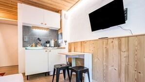 Cocina compacta con armarios blancos, encimera de madera y televisor integrado. Hay dos taburetes y una mesa pequeña.