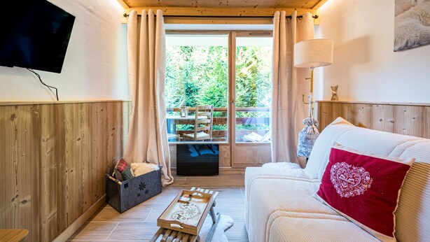 Una habitación acogedora con paredes de madera, un sofá blanco, un cojín rojo, un televisor montado en la pared y una ventana con cortinas.