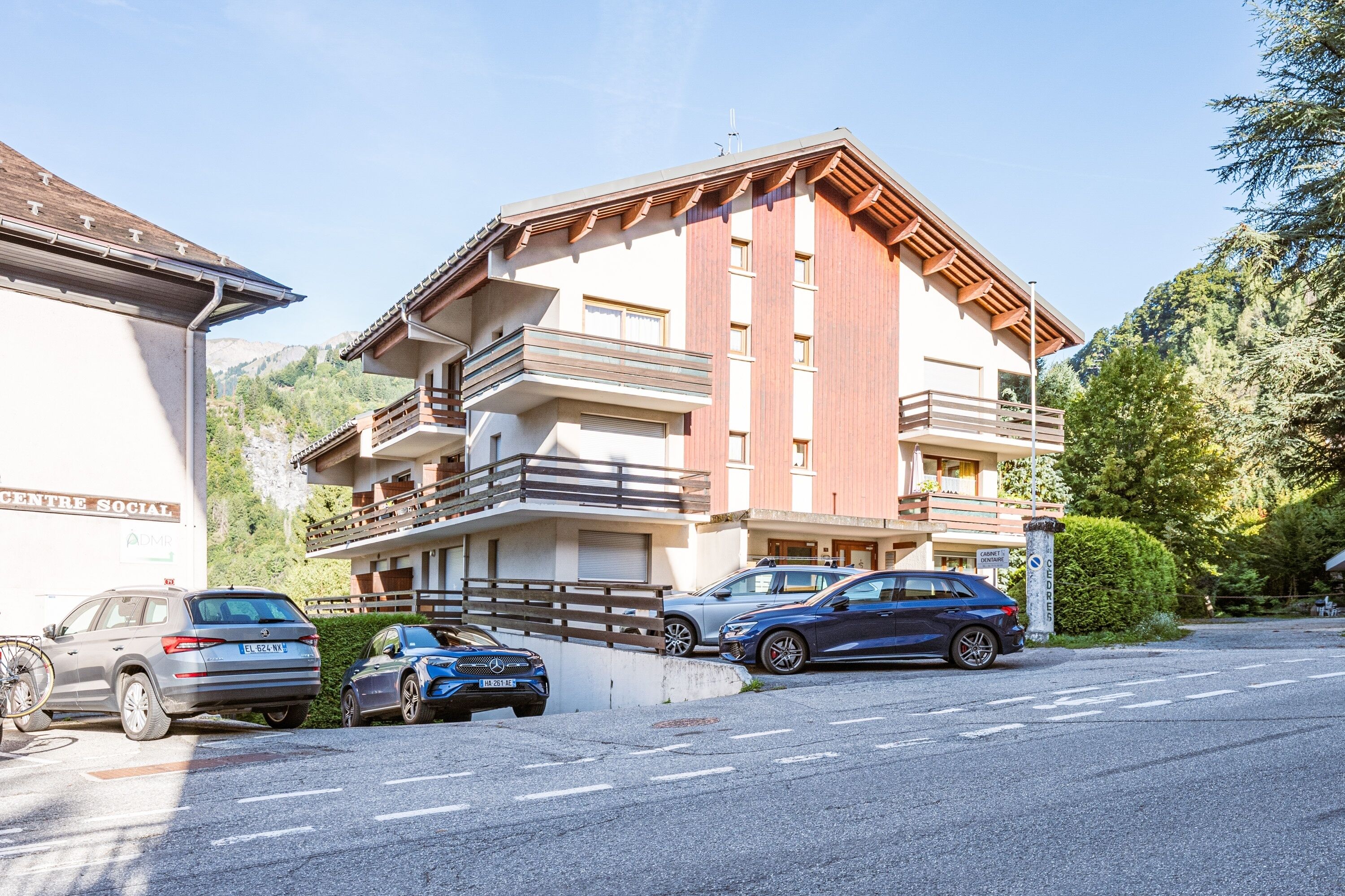 Un immeuble à plusieurs étages doté de balcons, un espace de stationnement avec plusieurs voitures et un paysage montagneux en arrière-plan.