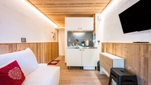 Habitación compacta con sofá blanco, pequeña área de cocina con microondas y hervidor, y televisor de pantalla plana montado en la pared.
