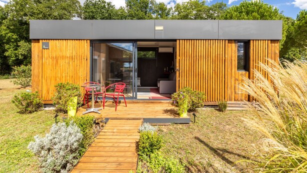 Uma cabana moderna de madeira com porta de entrada em vidro, uma pequena área de estar externa e um deck de madeira.