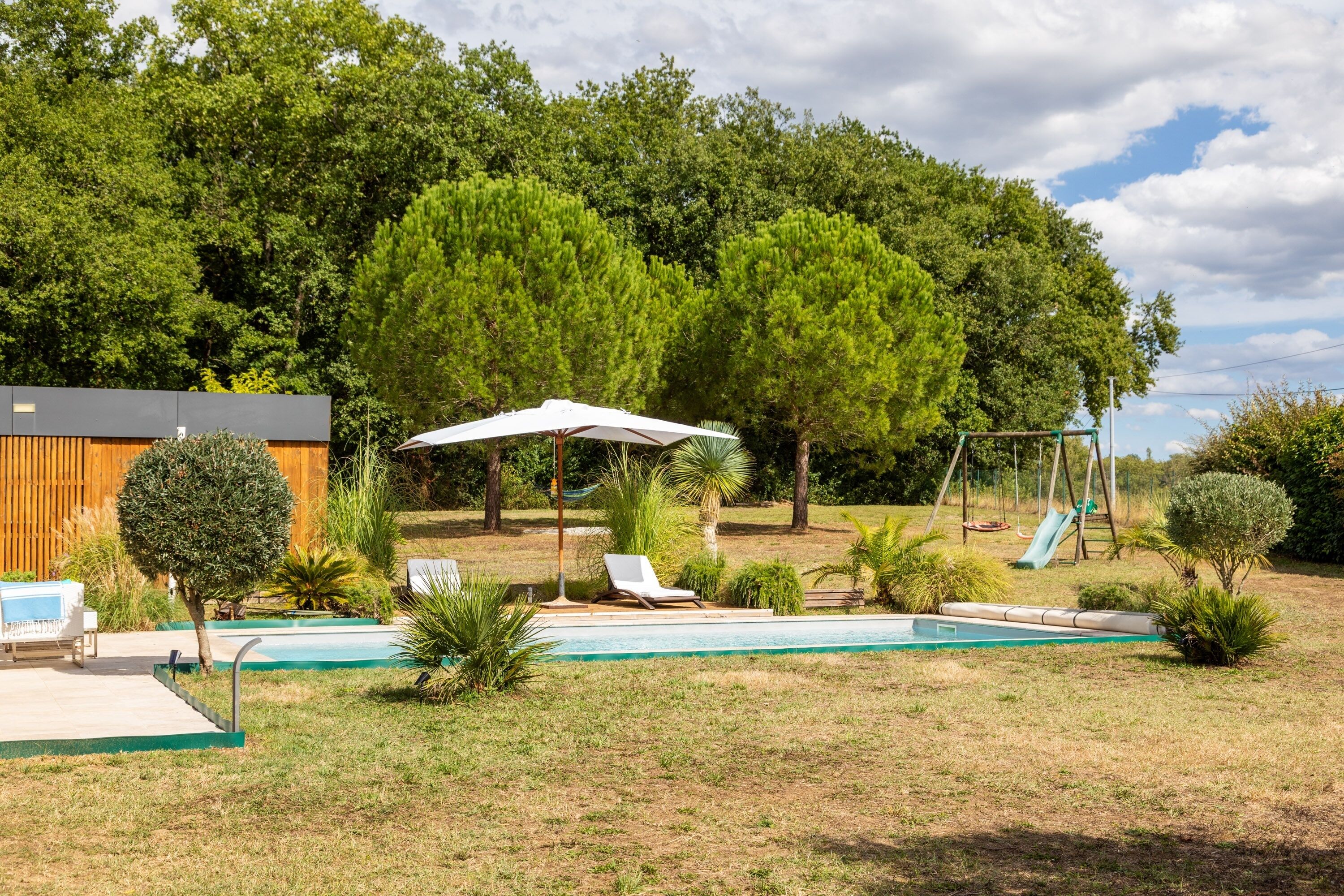 A backyard with a swimming pool, lounge chairs, a swing set, and a wooden house.
