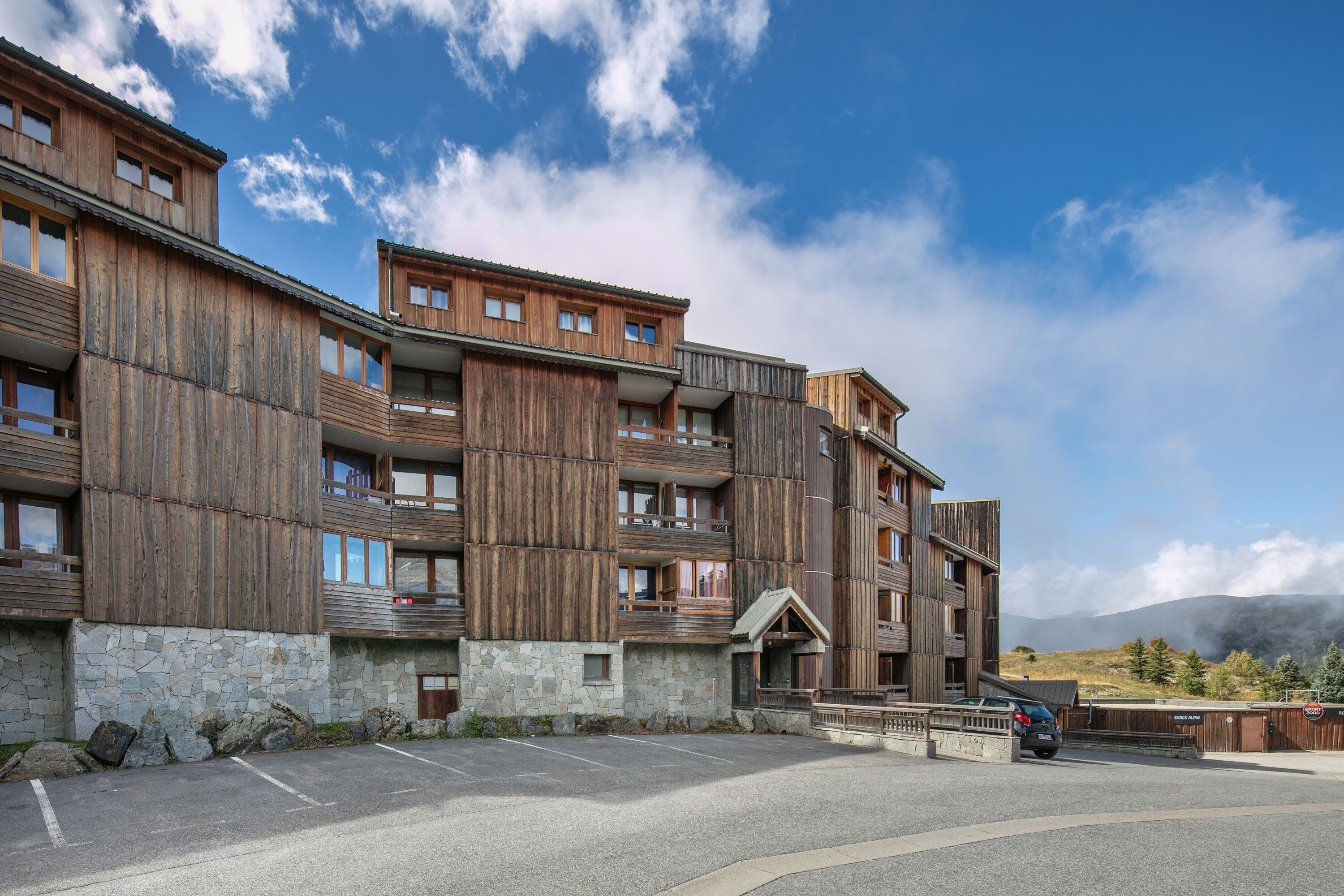 Un immeuble en bois à plusieurs étages, doté de balcons, d’une fondation en pierre et d’un espace de stationnement.