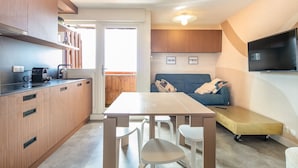Una cocina compacta con armarios de madera, una mesa de comedor con sillas, un sofá y un televisor de pantalla plana montado en la pared.