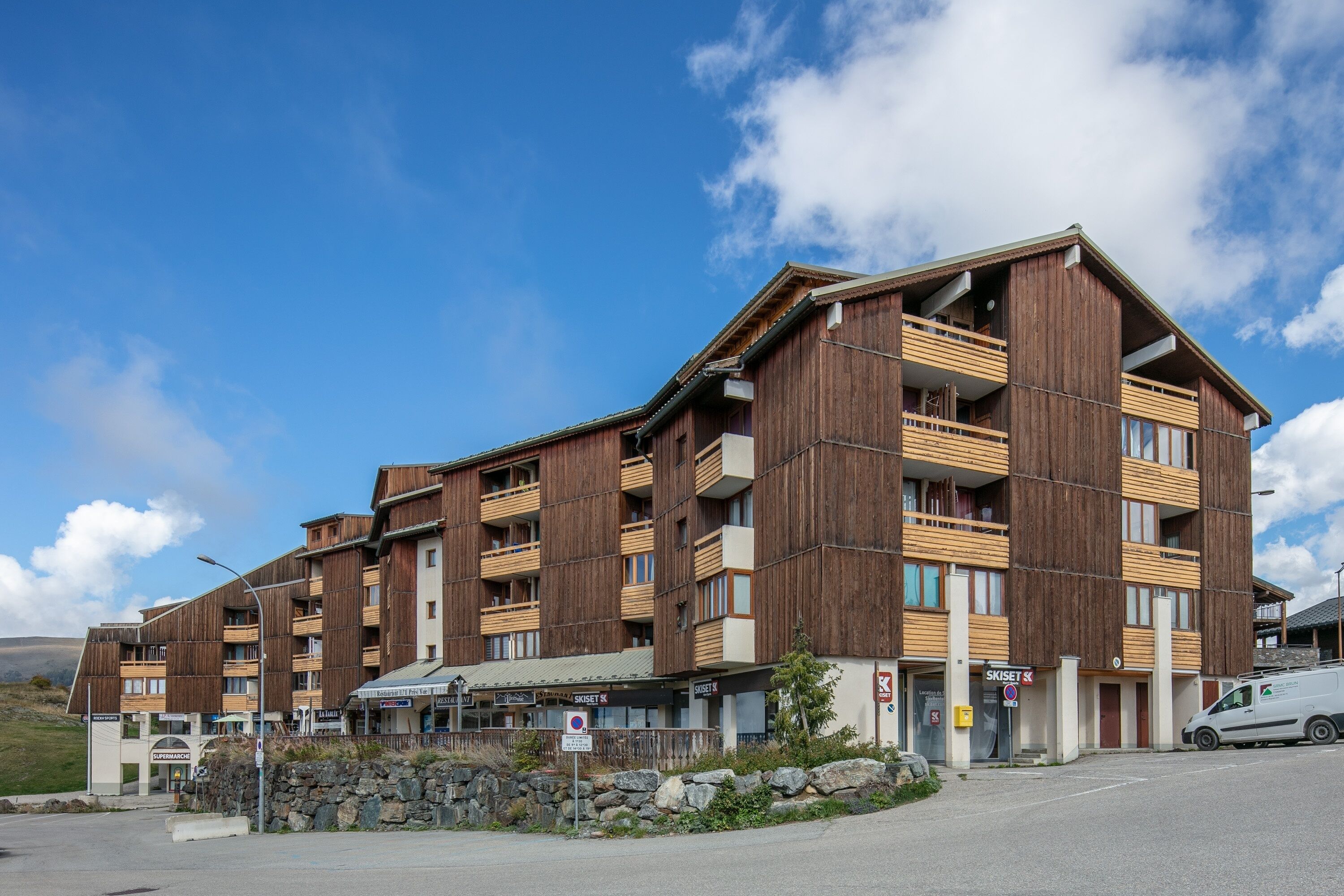 Un hôtel à plusieurs étages, doté d’une façade en bois et de balcons, situé près d’une route bordée d’un mur en pierre et d’un panneau indiquant qu’il est interdit de se garer.