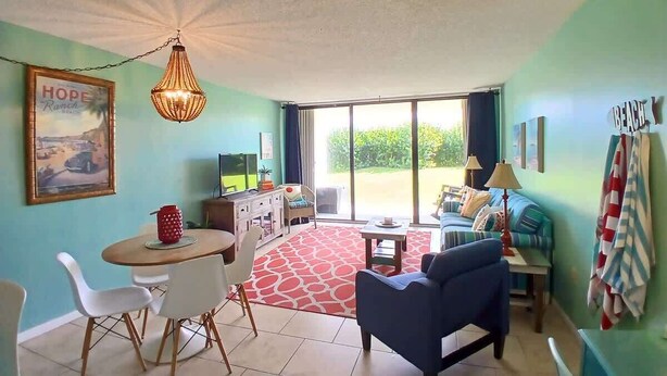 A living room with a dining area, a sofa, a TV, and a large window. - Aloha Awaits - 2B/2B Ground Floor Condo (Cape Canaveral)