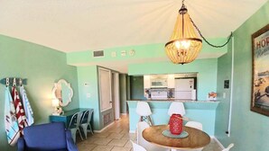 A dining area with a round table and chairs, a blue armchair, and a kitchen in the background. - Aloha Awaits - 2B/2B Ground Floor Condo (Cape Canaveral)