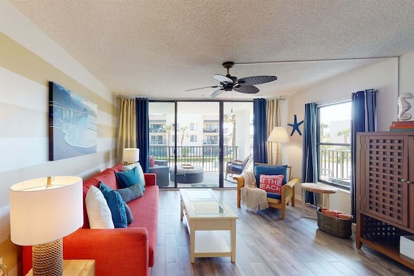 A modern living room with a red sofa, a coffee table, and a ceiling fan. There is a view of a balcony with a starfish decoration and a beach-themed pillow.
