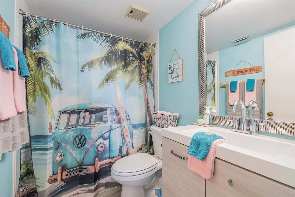 A bathroom with a tropical-themed shower curtain, a Volkswagen camper van mural, and a vanity with a mirror and towel rack.