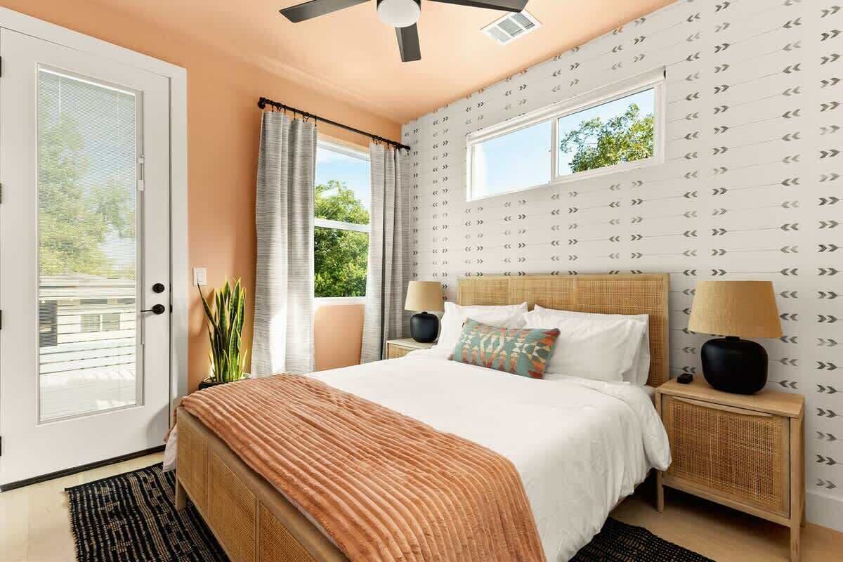 Ein Schlafzimmer mit einem Bett, zwei Nachttischen, einem Deckenventilator, einem Fenster mit Vorhängen und einer Tür.
