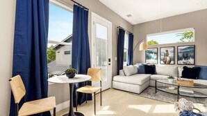 A modern living room with a white sofa, a glass coffee table, a round dining table with chairs, and large windows with blue curtains.