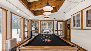 A pool table in a room with large windows, wooden beams on the ceiling, and a ceiling light fixture.