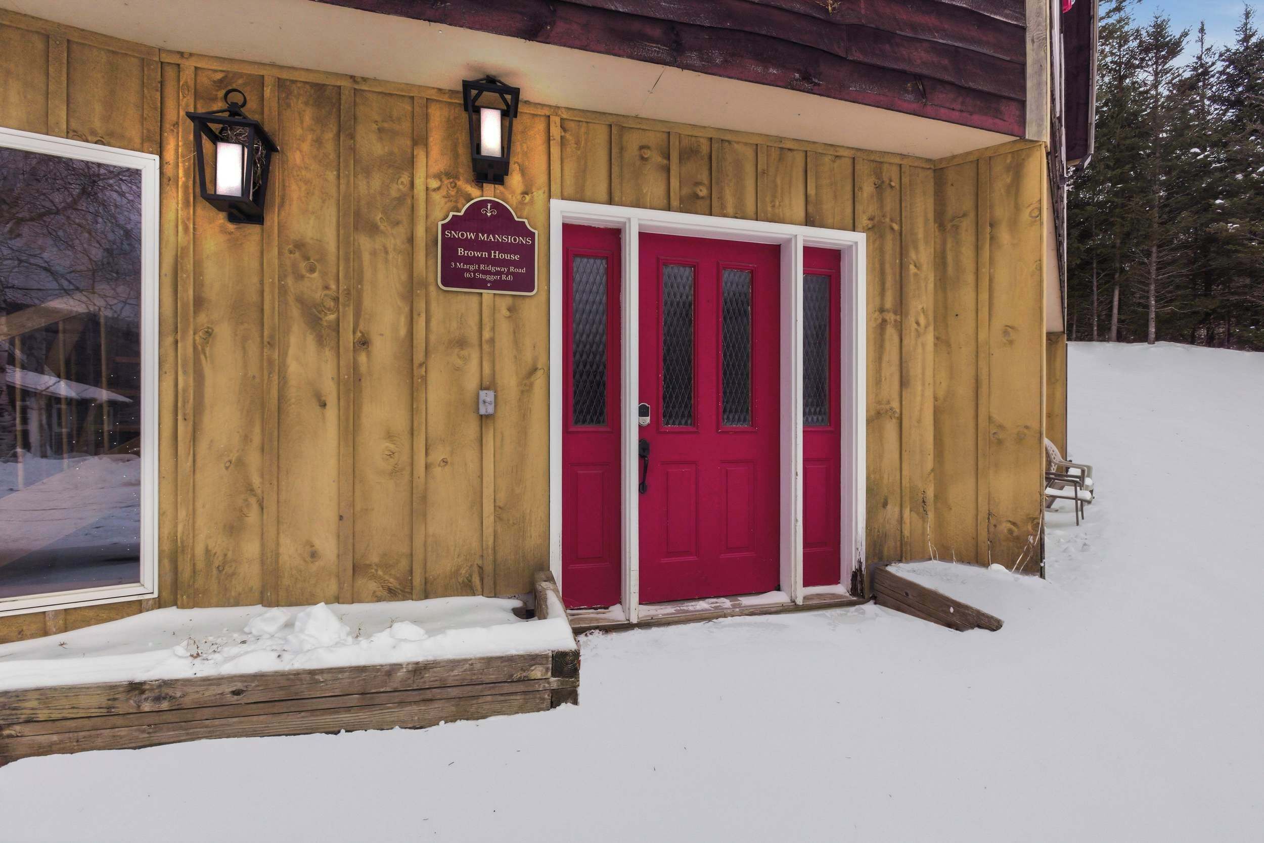 Une cabane en bois avec une porte rouge, une enseigne indiquant « Snow Mansions Brown House », et une fenêtre donnant sur un paysage enneigé.