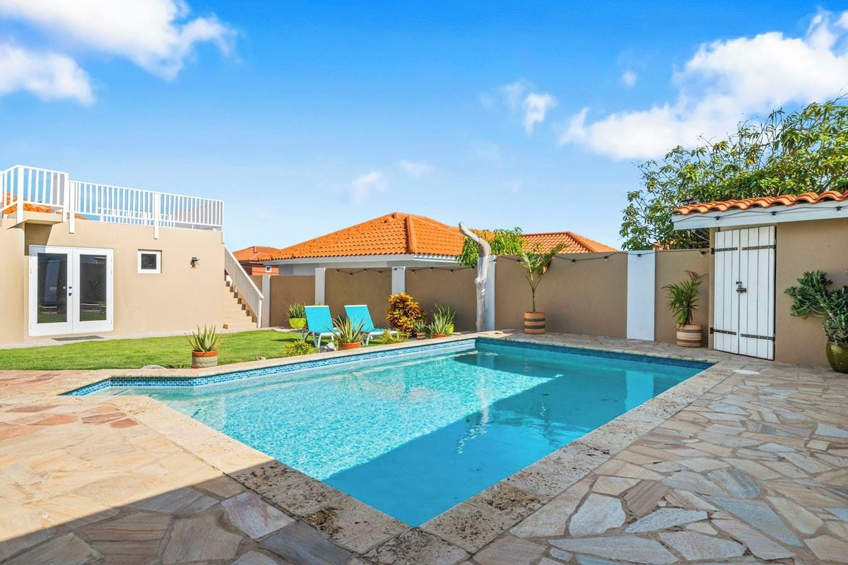 A swimming pool with a stone deck, a covered patio, and a two-story building with a red-tiled roof.