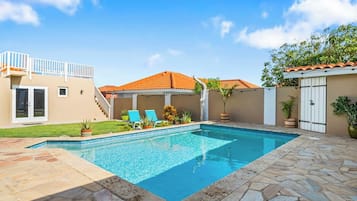 A swimming pool with a stone deck, a covered patio, and a two-story building with a red-tiled roof.