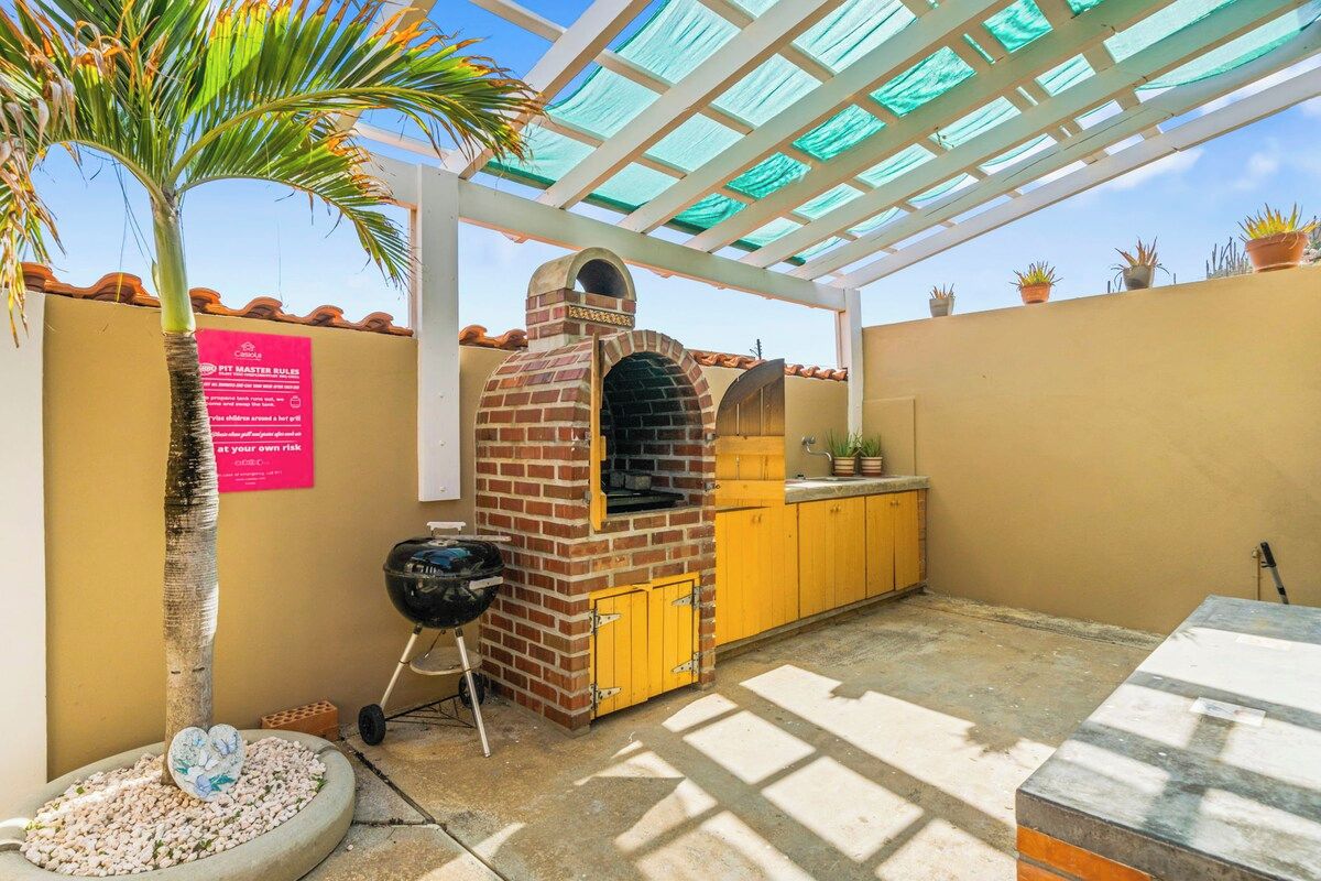An outdoor patio with a brick oven, yellow cabinets, a grill, and a palm tree.