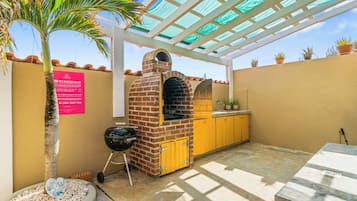 An outdoor patio with a brick oven, yellow cabinets, a grill, and a palm tree.