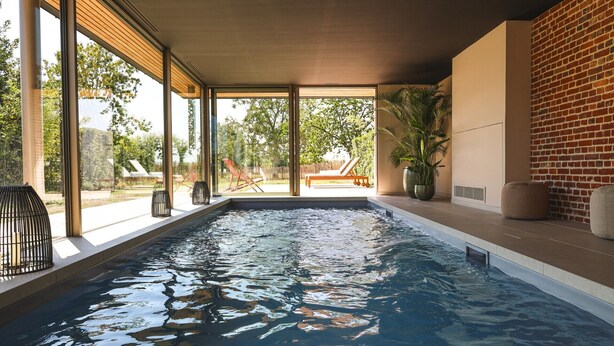 An indoor swimming pool with clear glass walls, a brick wall on one side, and a view of a garden with chairs and a palm tree. - Clos du Riez - Suites & SPA (La Gorgue)
