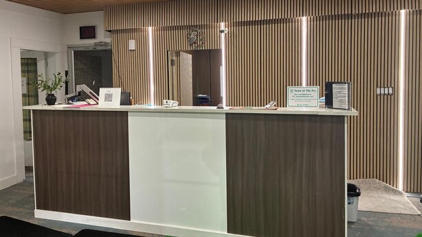 A reception desk with a modern design, featuring a wooden panel and a glass partition. There is a sign indicating the location as 'Town of The Pas' and a QR code on the desk. - The Pas Motel (The Pas)
