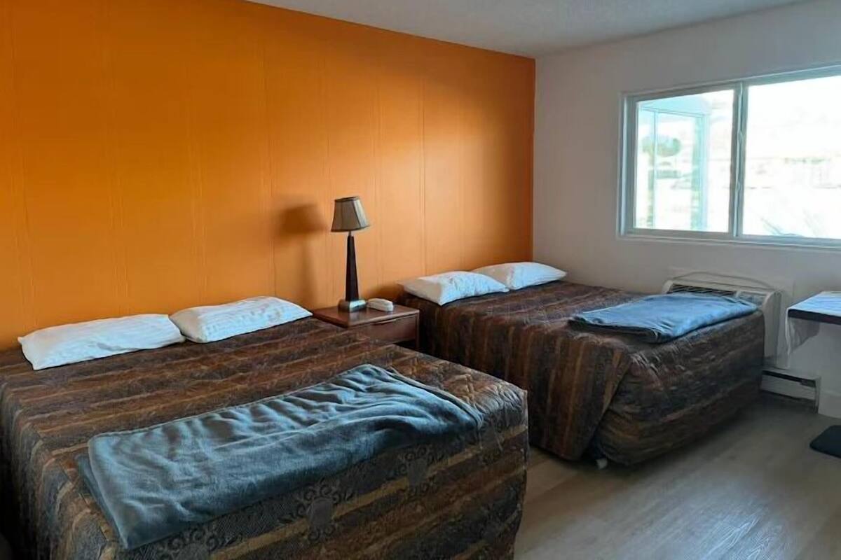 A hotel room with two beds, a nightstand, a lamp, and a window.
