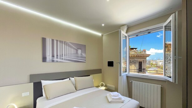 A hotel room with a neatly made bed, a bedside table with a lamp, a wall-mounted picture, and a window with a view of a building and sky.
