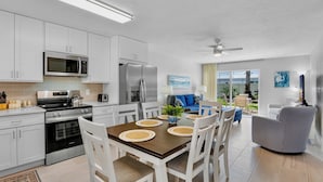Una cocina moderna con armarios blancos, electrodomésticos de acero inoxidable, una zona de comedor con mesa para cuatro personas y un área de estar con sofá y sillones.