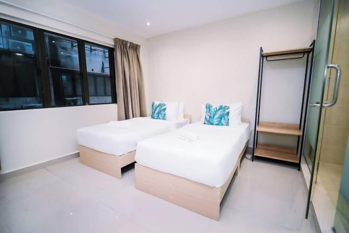 A hotel room with two beds, a window with curtains, a glass shower enclosure, and a wooden shelf.