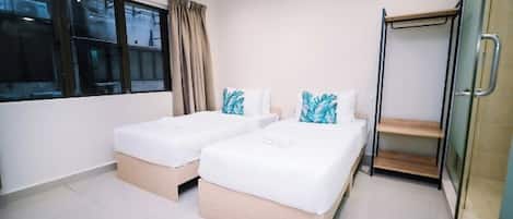 A hotel room with two beds, a window with curtains, a glass shower enclosure, and a wooden shelf.
