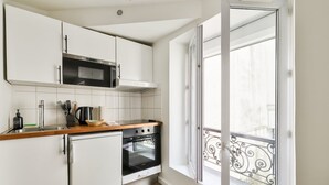 Cocina compacta con armarios blancos, microondas, estufa y un refrigerador pequeño. Hay una ventana con balcón exterior.