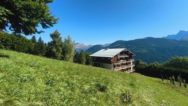 A wooden chalet with a balcony, surrounded by lush greenery and mountains in the background.