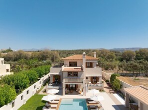 A two-story building with a swimming pool, surrounded by greenery and a clear sky.