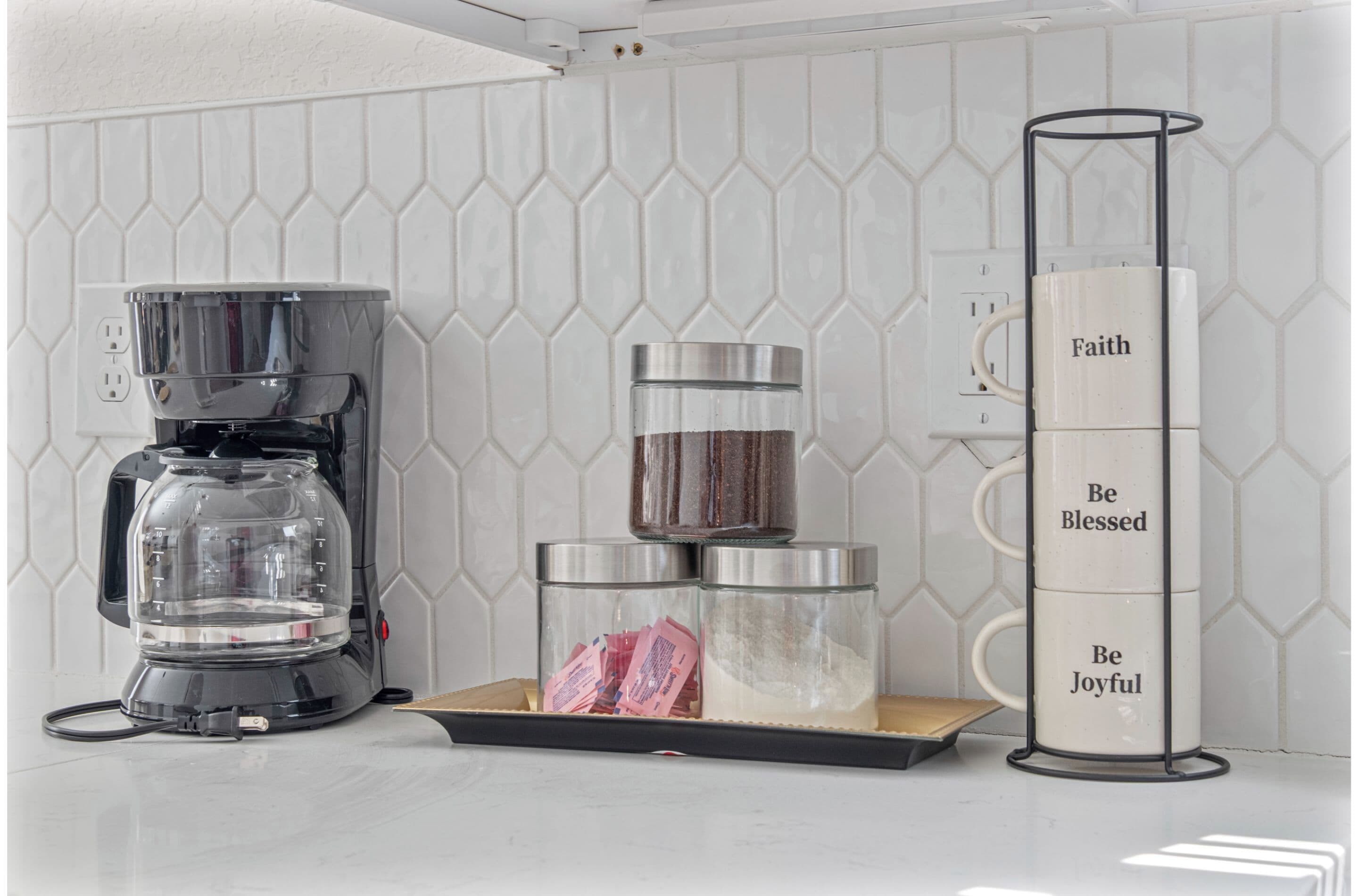 A kitchen counter with a coffee maker, two glass jars, and a mug stand with motivational phrases.