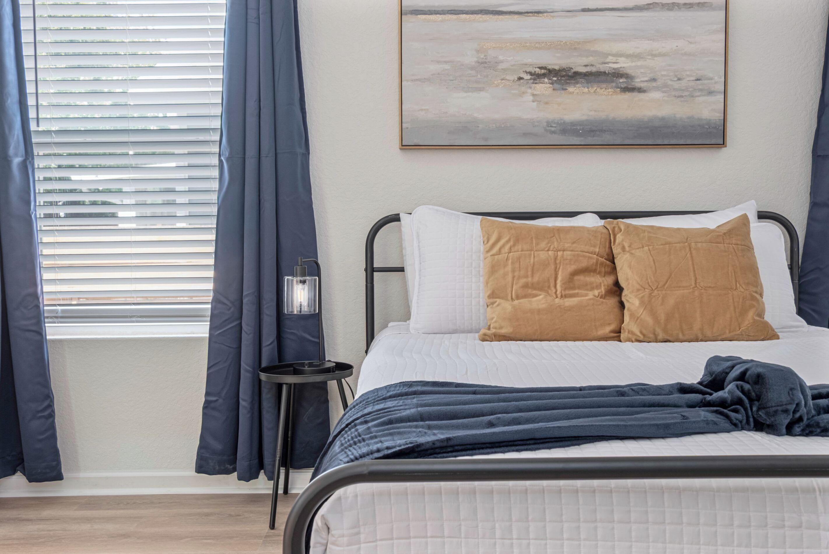 Una cama bien hecha con almohadas blancas y marrones, una manta azul oscuro y una mesita de noche con una lámpara.