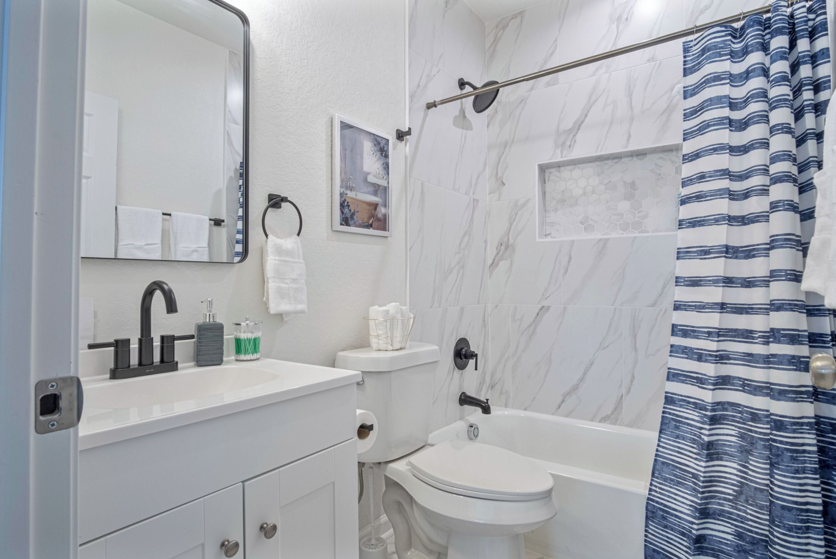 Une salle de bain avec un lavabo, des toilettes et une baignoire, tous de couleur blanche. On y trouve un rideau de douche, un porte-serviettes et un miroir.