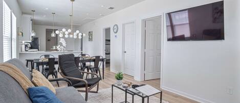 A modern living room with a gray sofa, black chairs, and a coffee table. There is a dining area with a table and chairs, and a kitchen in the background.