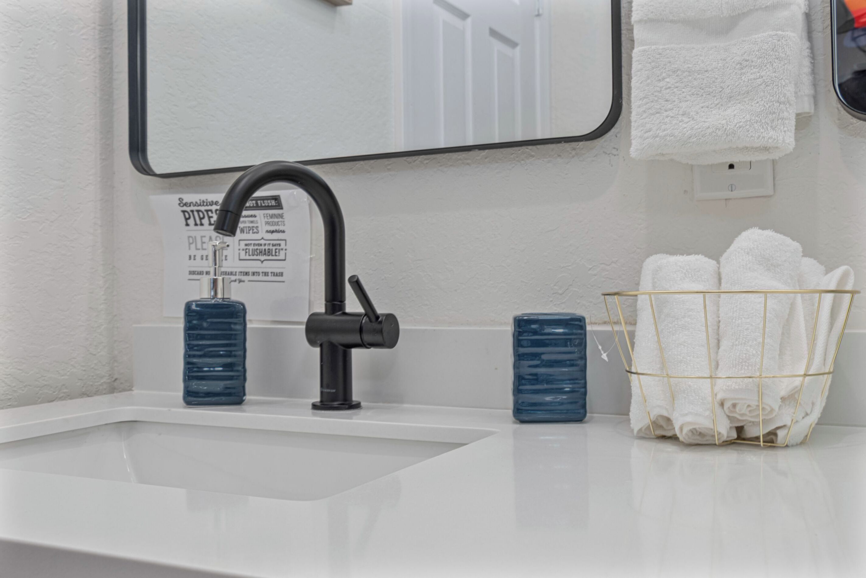 Un lavabo da bagno con un rubinetto nero, uno specchio sopra e un portasciugamani con asciugamani bianchi. Sul mobile due bottiglie azzurre.
