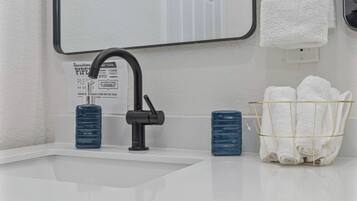 Un lavabo da bagno con un rubinetto nero, uno specchio sopra e un portasciugamani con asciugamani bianchi. Sul mobile due bottiglie azzurre.