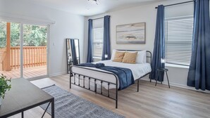 A bedroom with a bed, a ceiling fan, a window with curtains, a small table, and a view of a wooden deck.