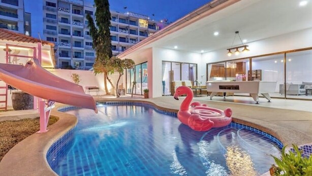 A swimming pool with a slide and a flamingo float, surrounded by a modern building with large windows and a well-lit interior. - 3 beds Pratamnak Near Walking Street (Chon Buri)